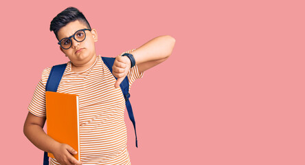 Little boy kid wearing student backpack holding books with angry face, negative sign showing dislike with thumbs down, rejection concept