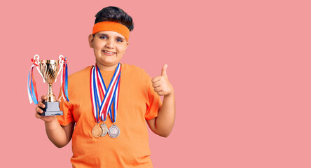 Little boy kid holding champion trophy and wearing medals smiling happy and positive, thumb up doing excellent and approval sign