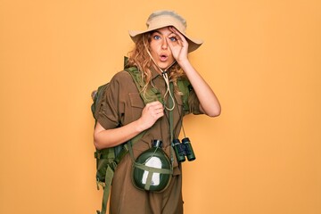 Young blonde explorer woman with blue eyes hiking wearing backpack and water canteen doing ok gesture shocked with surprised face, eye looking through fingers. Unbelieving expression.