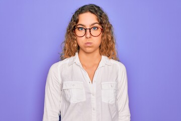 Young beautiful woman with blue eyes wearing casual shirt and glasses over purple background puffing cheeks with funny face. Mouth inflated with air, crazy expression.