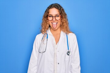 Young beautiful blonde doctor woman with blue eyes wearing coat and stethoscope sticking tongue out happy with funny expression. Emotion concept.