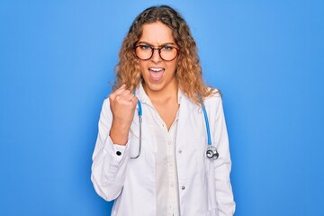 Young beautiful blonde doctor woman with blue eyes wearing coat and stethoscope angry and mad raising fist frustrated and furious while shouting with anger. Rage and aggressive concept.