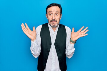 Middle age handsome man wearing elegant shirt and vest over isolated blue background clueless and confused with open arms, no idea and doubtful face.