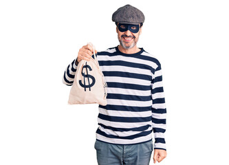 Middle age handsome man wearing burglar mask holding money bag looking positive and happy standing and smiling with a confident smile showing teeth