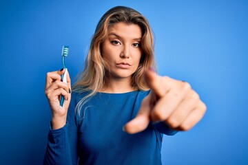 Young beautiful blonde woman using teeth brush over isolated blue background pointing with finger to the camera and to you, hand sign, positive and confident gesture from the front