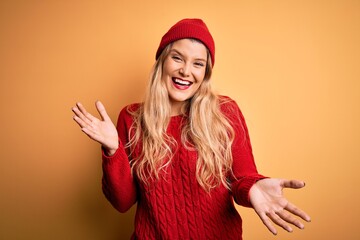 Young beautiful blonde woman wearing casual sweater and wool cap over white background smiling cheerful with open arms as friendly welcome, positive and confident greetings