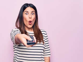 Young beautiful brunette woman holding television remote control over pink background scared and amazed with open mouth for surprise, disbelief face