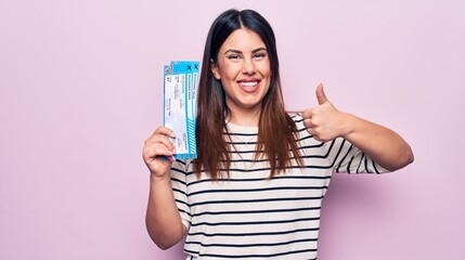 Young beautiful tourist woman holding airline boarding pass over isolated pink background smiling happy and positive, thumb up doing excellent and approval sign