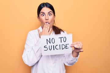 Young beautiful brunette psychologist woman holding paper with not to suicide message covering mouth with hand, shocked and afraid for mistake. Surprised expression