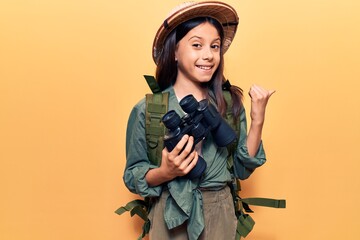 Beautiful child girl wearing explorer hat holding binoculars pointing thumb up to the side smiling happy with open mouth