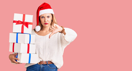Fototapeta premium Beautiful young woman wearing christmas hat and holding a gift with angry face, negative sign showing dislike with thumbs down, rejection concept