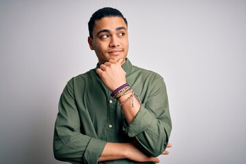 Young brazilian man wearing casual shirt standing over isolated white background with hand on chin thinking about question, pensive expression. Smiling with thoughtful face. Doubt concept.