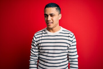 Young brazilian man wearing casual striped t-shirt standing over isolated red background winking...