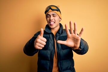 Young brazilian skier man wearing snow sportswear and ski goggles over yellow background showing and pointing up with fingers number six while smiling confident and happy.
