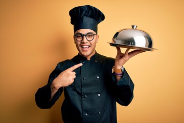 Young brazilian chef man wearing cooker uniform and hat holding tray with dome very happy pointing with hand and finger