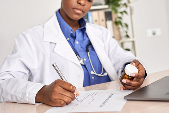 African american female medic doctor gp therapist wear white coat holding pills bottle in hand writing medical prescription sitting at work desk prescribing pharmacy medicine concept. Closeup view