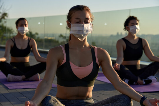 Fit sporty young woman wear medical face mask for safety meditating keeping social distance with female friends practicing yoga fitness exercise together on sunrise at retreat group class outdoors.