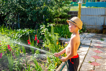 a boy in a hat watering flowers from a hose on a hot summer day