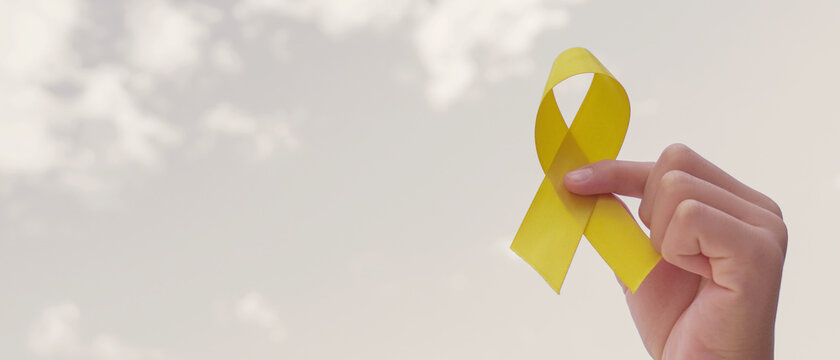 Hands holding yellow gold ribbon over grey sky, Sarcoma Awareness, Bone cancer, childhood cancer awareness, September yellow, World Suicide Prevention Day concept