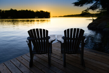 Fototapeta na wymiar Silhouette of two Muskoka chairs sitting on a cottage wooden dock at sunset. The warm orange colours are contrasting with the blue sky. 