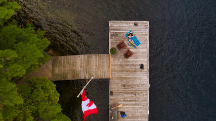From above, a wooden dock graces the serene Muskoka lakefront, where a young woman relaxes on a...