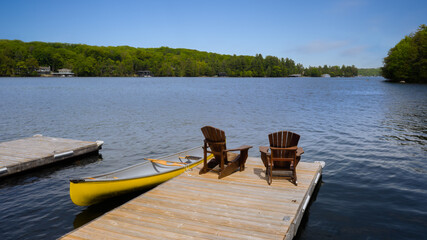 Fototapeta premium Two Adirondack chairs on a wooden dock facing a calm lake in Muskoka, Ontario Canada. A yellow canoe is tied to the dock. A cottage nestled between trees is visible across the water.