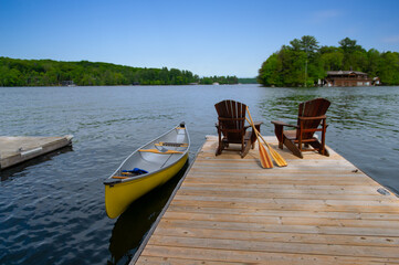 Fototapeta premium Two Adirondack chairs on a wooden dock facing the blue water of a lake in Muskoka, Ontario Canada. A yellow canoe is tied to the dock while the paddles are next to the chairs.