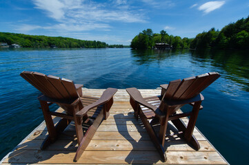 Close up of two Adirondack chairs sitting on a wooden dock facing the calm water of a lake in Muskoka, Ontario Canada. A cottages nestled between trees is visible in background. 