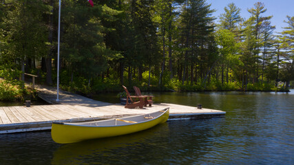 Two Adirondack chairs on a wooden dock facing a calm lake in Muskoka, Ontario Canada. A yellow canoe is tied to the dock. A cottage nestled between trees is visible across the water.