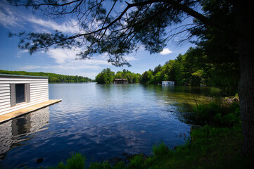 Lake view during a summer day at the cottage in Muskoka, Ontario Canada. A white boathouse building is visible. Cottages are nestled between trees along the shores.