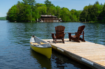 Fototapeta na wymiar Two Adirondack chairs on a wooden dock facing a calm lake in Muskoka, Ontario Canada. A yellow canoe is tied to the dock. A cottage nestled between trees is visible across the water.