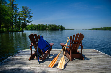 Two Adirondack chairs on a wooden dock facing the blue water of a lake in Muskoka, Ontario Canada....