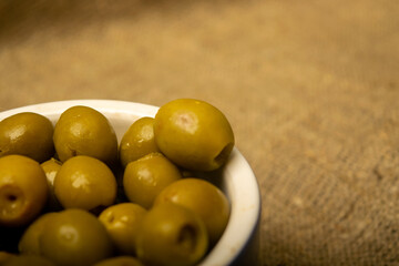 Green olives in a ceramic bowl on a background of coarse-textured burlap. Close up.