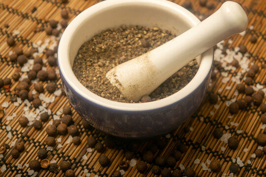 Ground black allspice in a ceramic mortar and black allspice and coarse salt scattered on a reed Mat. Close up.