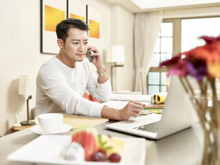 young asian business man working from home talking on cellphone while looking at laptop