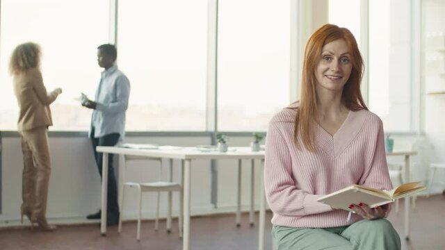 Portrait shot footage of beautiful mature woman with long red hair sitting on chair in office room holding copybook and looking it through