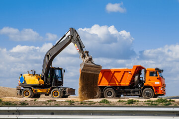 A yellow backhoe loader loads the earth into a truck during the construction of a road against a...