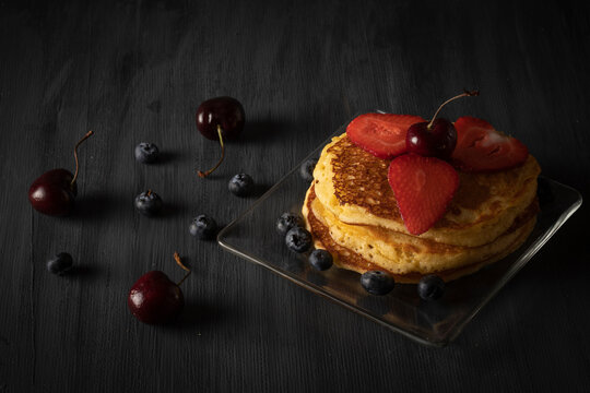Hot Cakes apilados con cerezas fresas y arandanos sobre una mesa de madera color negro vintage. Espacio para texto 