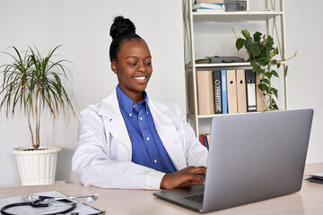 Smiling african american female doctor gp wears white medical coat using laptop computer at...