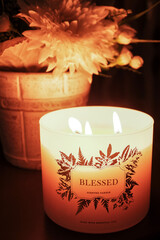 Lit scented candle with "Blessed" text, pot of flowers in the background