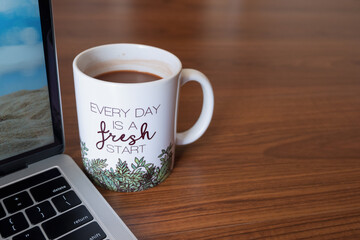 Coffee mug with inspirational message beside laptop on wooden surface. Every day is a fresh start. Copy space.