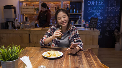Cheerful asian chinese girl drinking bottle of beer and eating chips. charming lady sitting at bar table in midnight alone using smart phone. happy young woman smiling looking at cellphone screen.