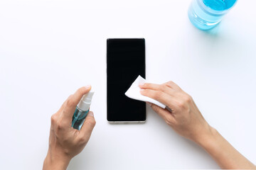 Woman hands cleaning smartphone with disinfectant wet wipe and alcohol spray. Concept of disinfecting daily use item from bacteria or viruses. Top view