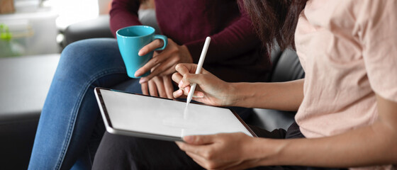 Two female sitting on black sofa, red shirt woman holding cup and pink shirt woman using digital tablet