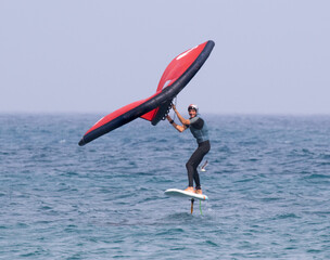 A man is wing foiling using handheld inflatable wings and hydrofoil surfboards in a blue ocean, this is a new wind sport that is becoming very popular quickly.
