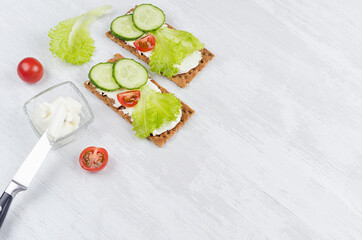 Fresh spring vitamine sandwiches of flat cereal rye dry crisps bread with green lettuce, cucumber, tomato, cream cheese on white wooden table, top view, copy space.
