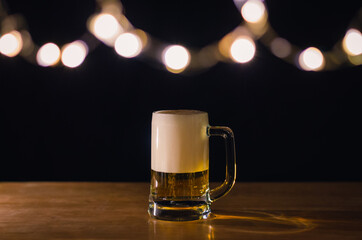 A glass of beer on wooden table that have bokeh lights on top with dark background.