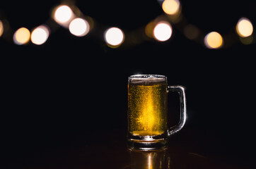 A glass of beer on wooden table that have bokeh lights on top with dark background.