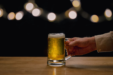 Hand holding a glass of beer on wooden table that have bokeh lights on top with dark background.