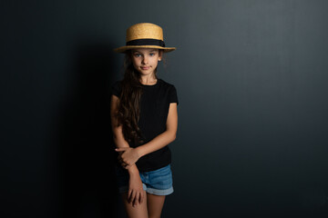 Beautiful caucasian little girl wearing black t0shirt and straw hat posing over deep gray wall looking at camera. Fashion portrait of cute kid.